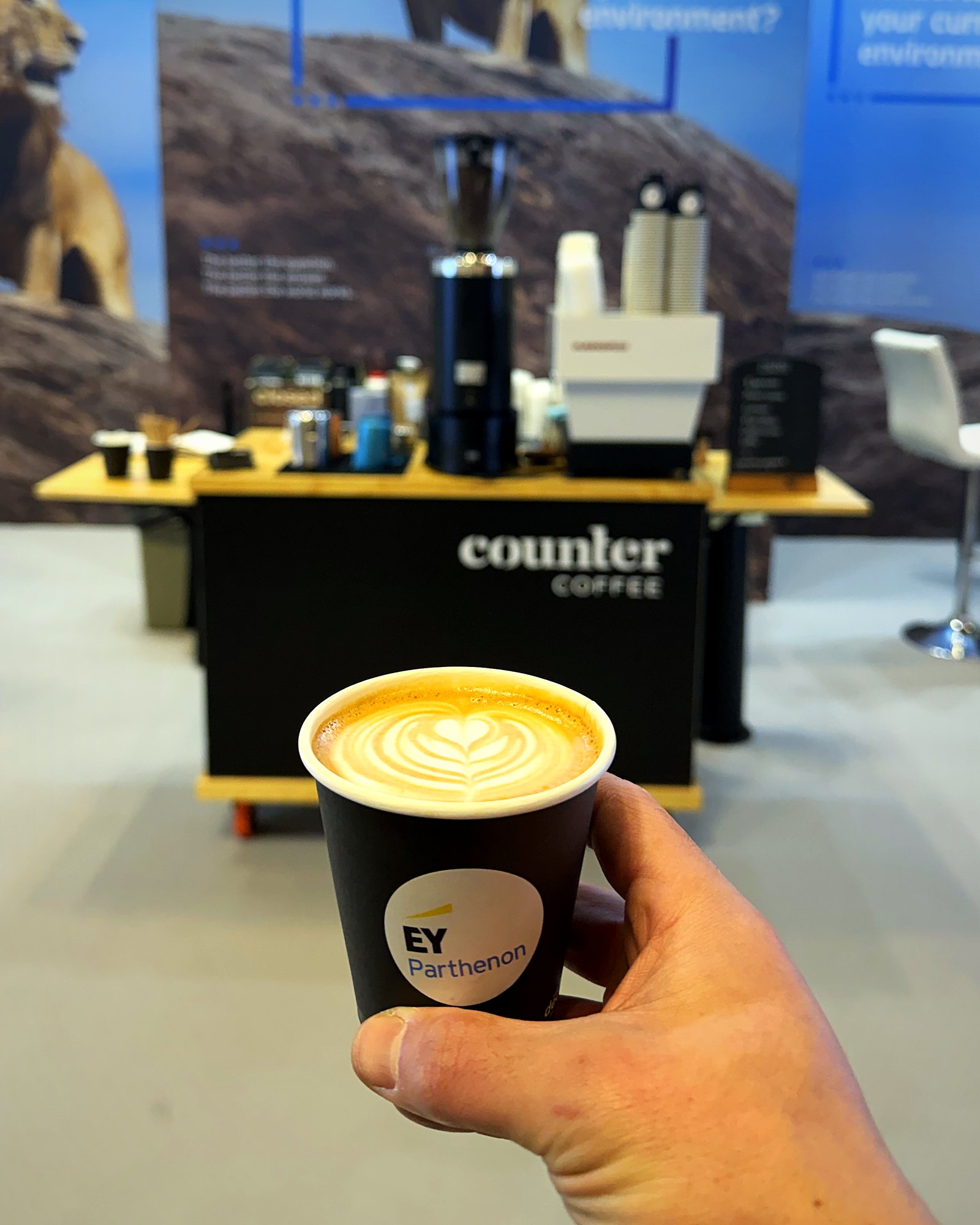 A latte in an EY Parthenon cup held up in front of the Counter Coffee cart at an EY conference